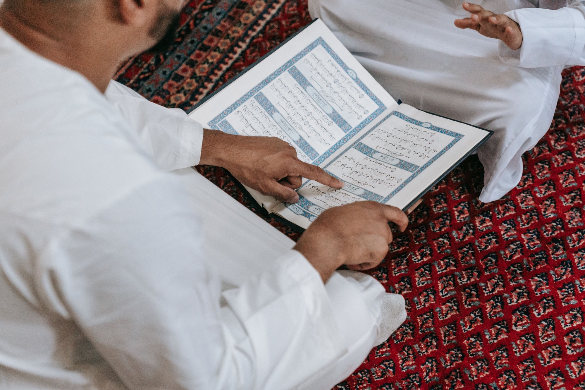 Two men seated on a red mosque carpet reading a Qur'an together