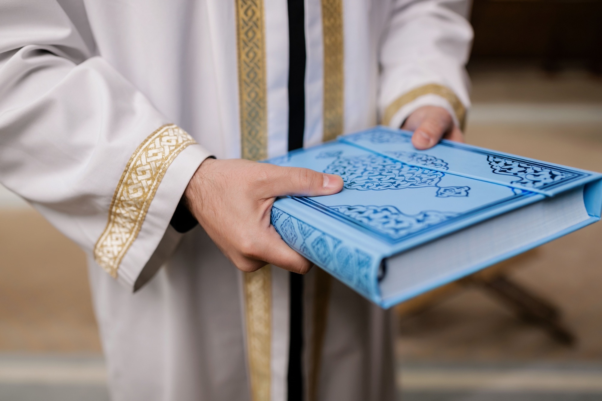 A man in a white thobe holding a closed mushaf, about to open it again