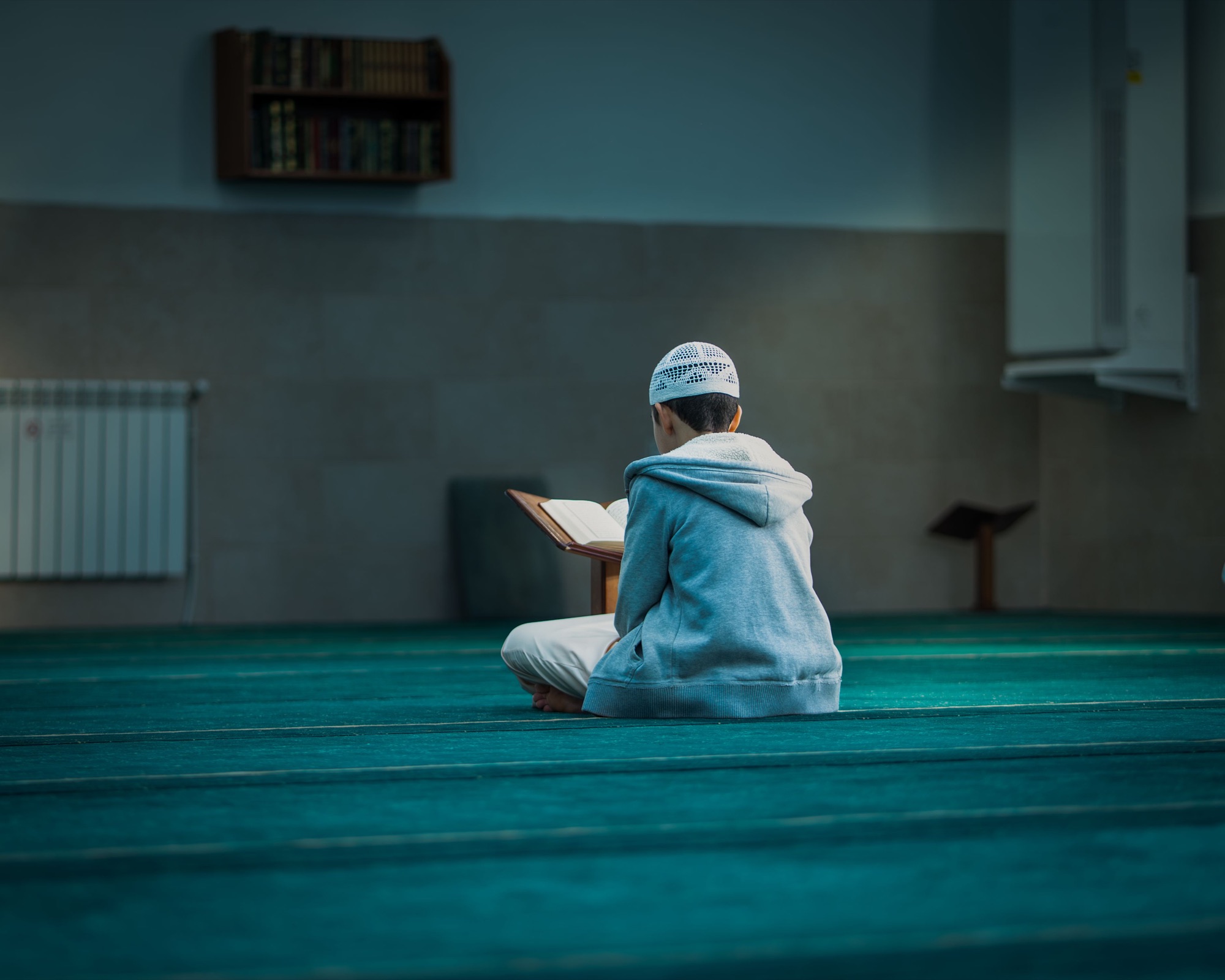 A boy sitting alone in a masjid, reading from a mushaf on a wooden stand