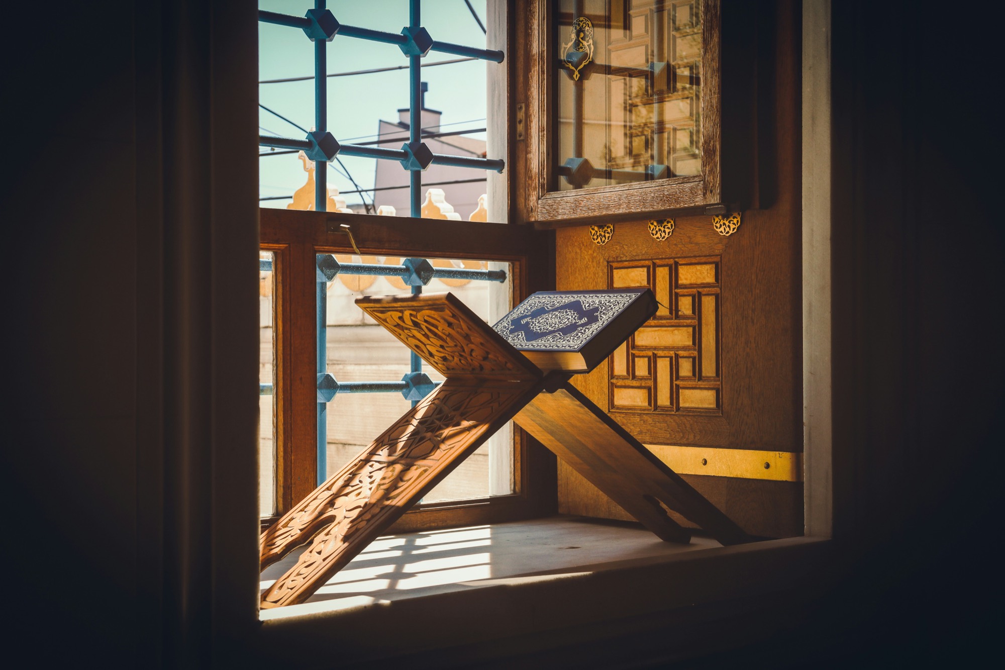 A carved wooden rehal holding an open mushaf in a sunlit window alcove
