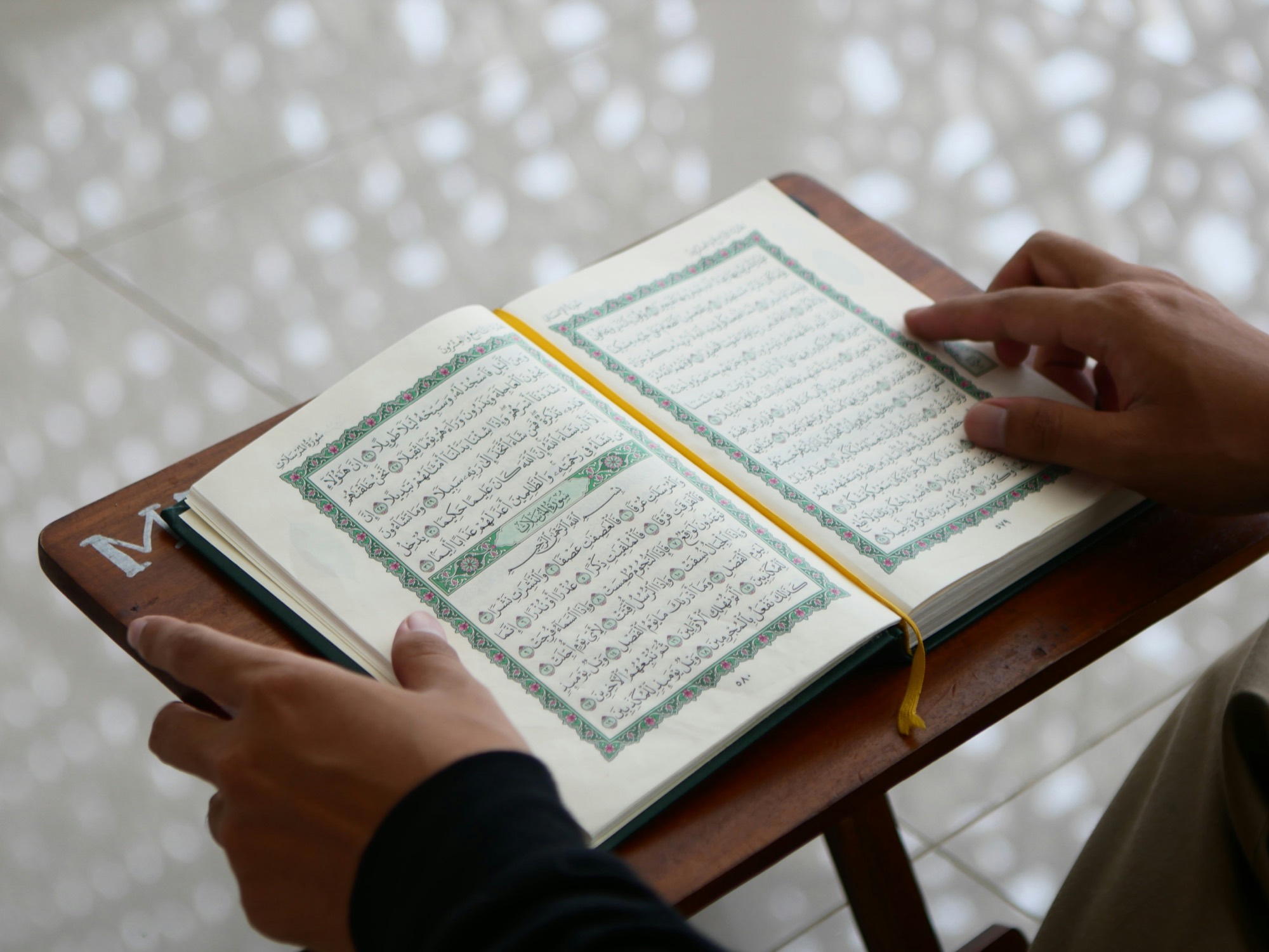 Hands resting on an open mushaf held on a small wooden folding stand