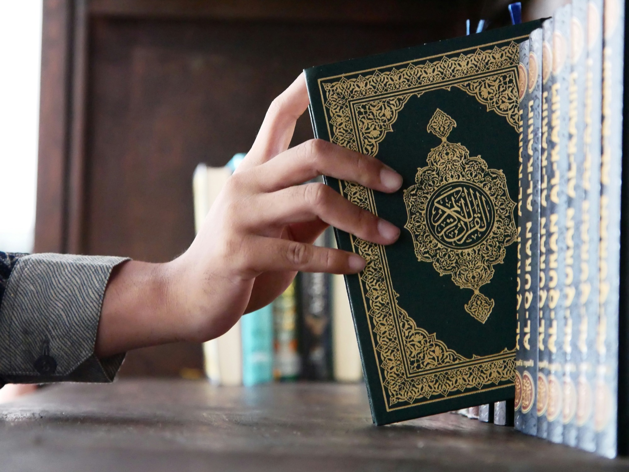 A hand reaching to pull a green mushaf from a row of copies on a shelf