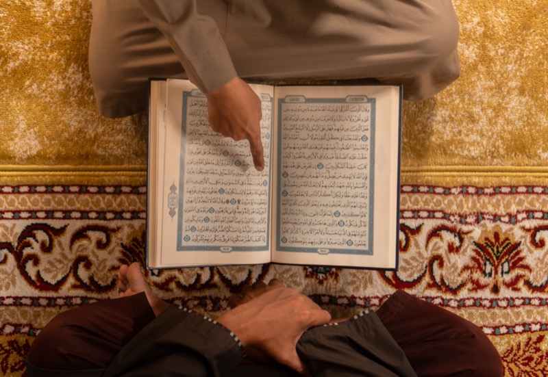 A man seated on a patterned mosque carpet reading an open Qur'an