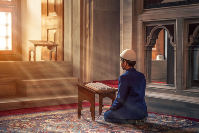 A young boy sitting before a Qur'an on a rehal in a sunlit mosque