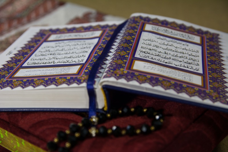 An ornately bordered Qur'an open at Surah Al-Fatihah with prayer beads