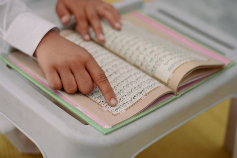 A child's hand tracing the lines of an open Qur'an on a desk
