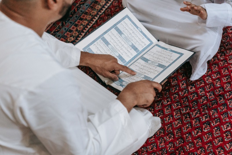 Two men seated on a red mosque carpet reading a Qur'an together