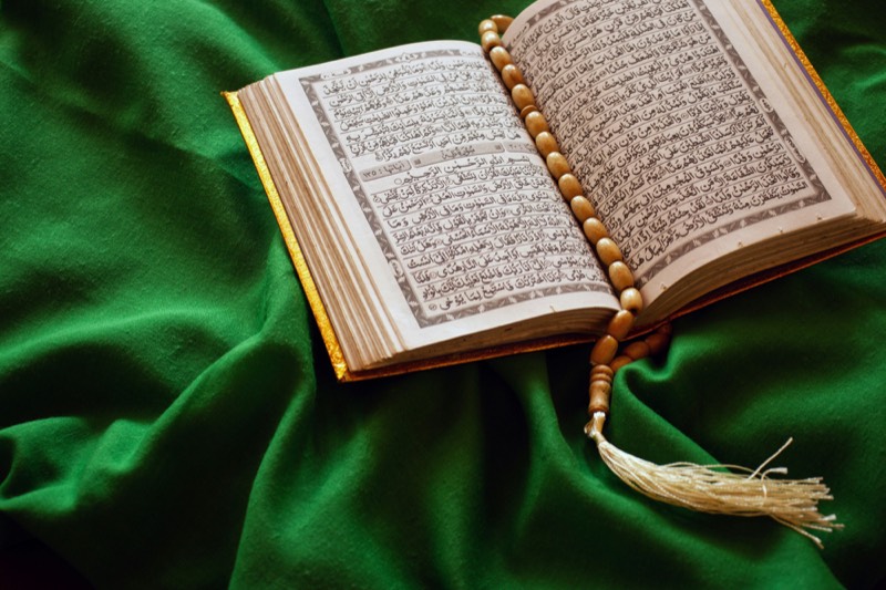 An open mushaf with a wooden tasbih resting across its pages on green cloth
