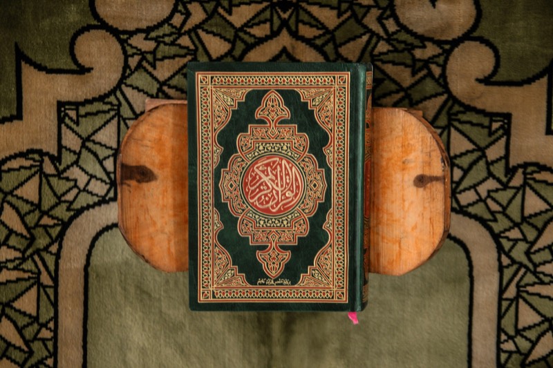 A green and gold mushaf standing closed on a wooden rehal over a prayer rug