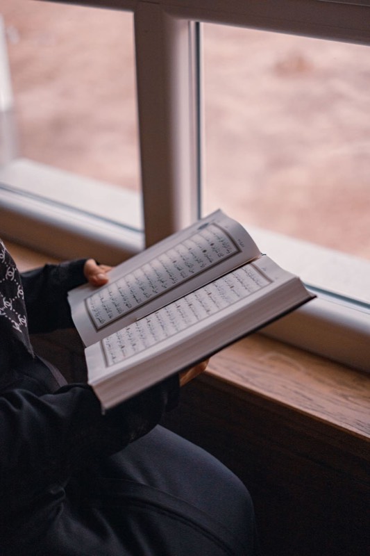 A person holding an open mushaf by a window, reciting quietly